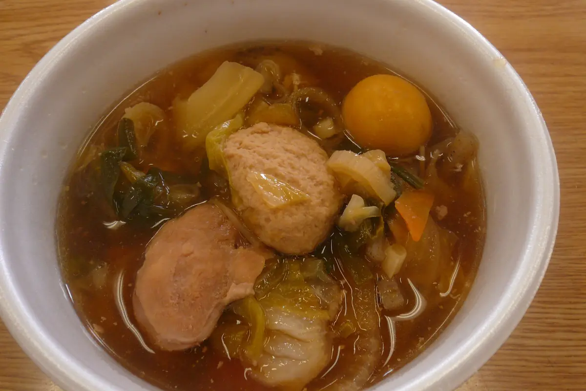 A white bowl of chankonabe stew with a fish ball, an egg yolk, cabbage and carrot in a brown broth, on a wooden table