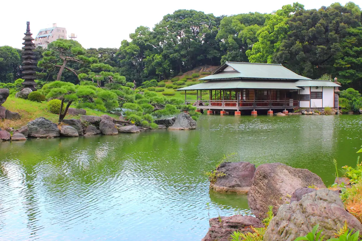 A pond with a tea house on a wooded island, rocks along the shore and a stone pagoda among pine trees in a Japanese garden