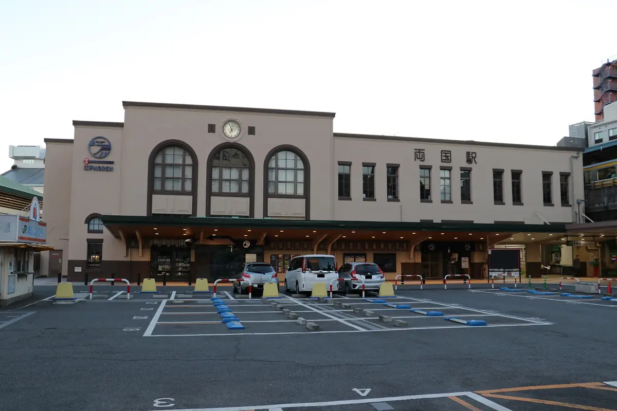 The cream-coloured main building of Ryogoku station with arched windows and a clock, and a car park in front