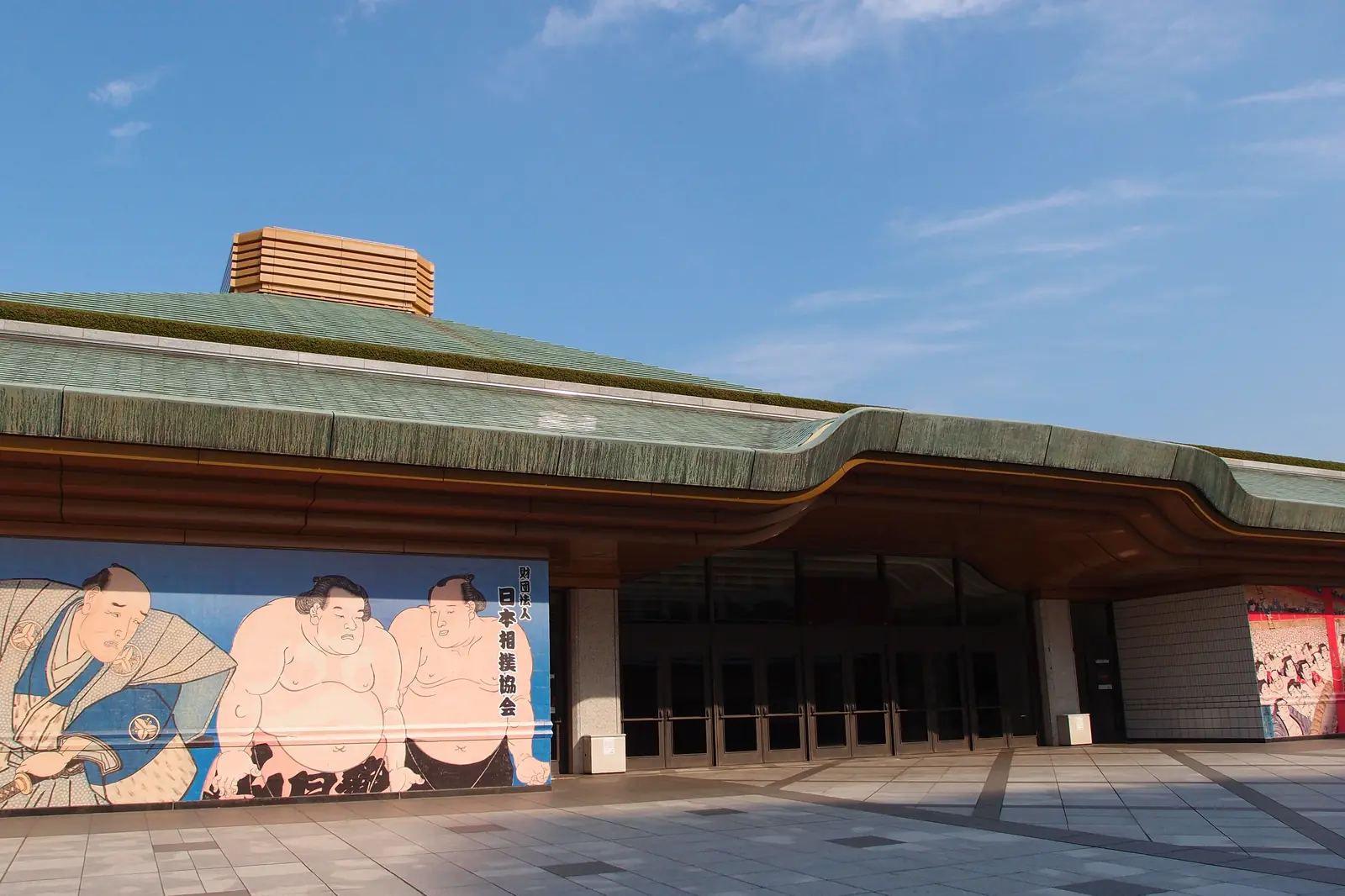 Ryogoku Kokugikan, Tokyo's sumo arena, seen from the front with its long green roof, wide entrance hall and a painted mural of wrestlers beside the doors.
