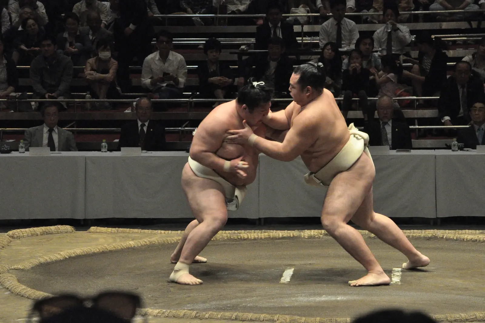 Two sumo wrestlers gripping each other on a clay ring at a practice session, with spectators in the stands behind and a judges' table beyond the ring