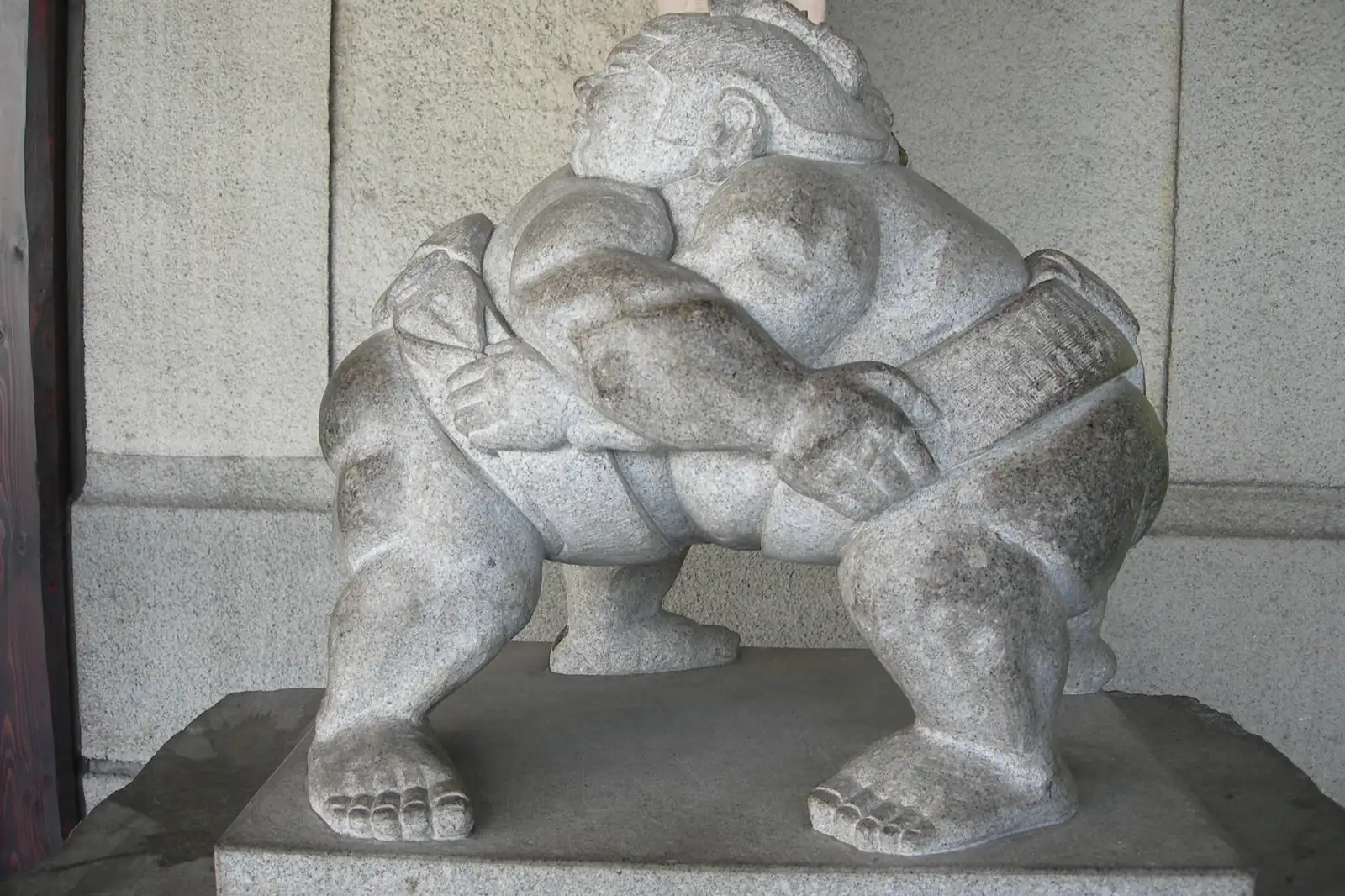 A grey stone statue of two sumo wrestlers locked in a hold, standing in a station entrance with a polished stone wall behind